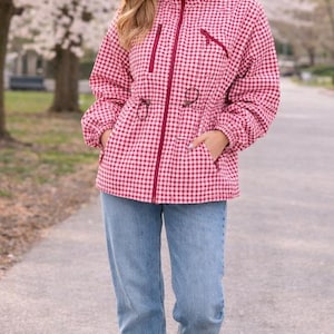 May include: A woman wearing a red and white gingham print hooded jacket with a drawstring waist, paired with light blue jeans and white sneakers. The jacket has a zipper closure and pockets. The background features a tree-lined path.