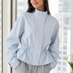 May include: A light blue jacket featuring a high collar, long sleeves with buttoned cuffs, and a cinched waist with a drawstring cord. The front closure includes a concealed placket with visible snap buttons, creating a structured silhouette.