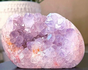 May include: A polished amethyst geode with a crystalline interior. The geode features a mix of light purple and pink hues, with visible crystal formations. The overall shape is rounded, showcasing the natural beauty of the gemstone.