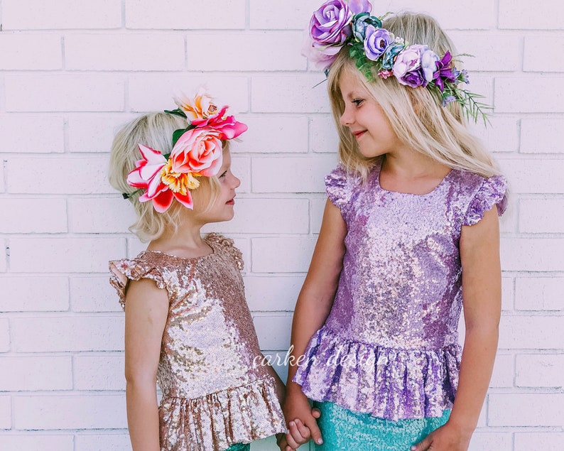 Puede incluir: Dos chicas con coronas de flores y tops de lentejuelas. La chica de la izquierda lleva una corona de flores rosa y blanca y un top de lentejuelas dorado. La chica de la derecha lleva una corona de flores moradas y un top de lentejuelas morado.