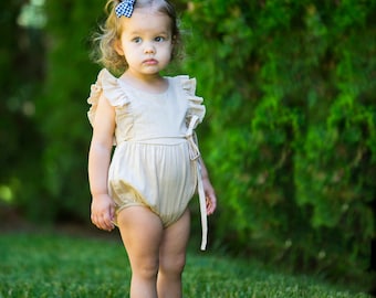 May include: A young girl wearing a light beige ruffled romper with a blue and white gingham bow in her hair. She is standing in a grassy area with green foliage in the background.