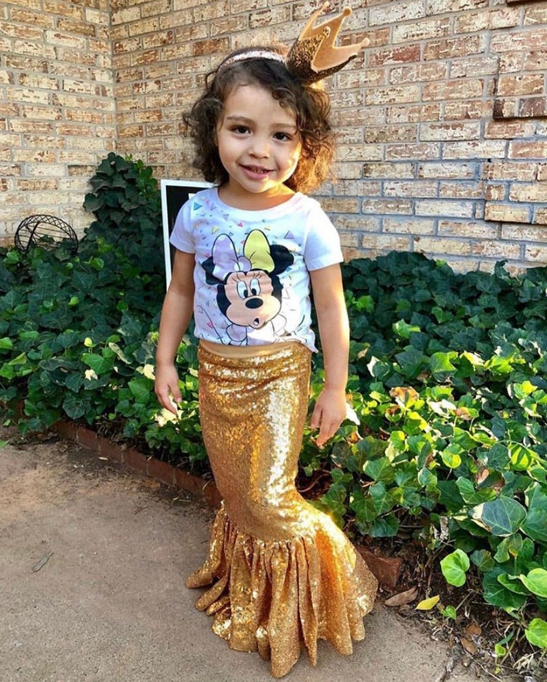 Może przedstawiać: Mała dziewczynka w białej koszulce z kresk&oacute;wkowym wizerunkiem Minnie Mouse i złotą, cekinową sp&oacute;dnicą syreny. Ma na głowie złotą, błyszczącą opaskę z koroną.
