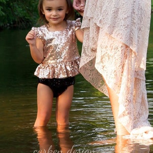 Puede incluir: Una ni&ntilde;a peque&ntilde;a con un top de lentejuelas de color oro rosa y una falda de volantes negra, con una flor blanca en el pelo, est&aacute; de pie en un arroyo de agua poco profundo. Est&aacute; cogida de la mano de un adulto que lleva una falda de encaje blanca.