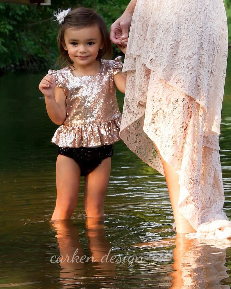 Puede incluir: Una ni&ntilde;a peque&ntilde;a con un top de lentejuelas de color oro rosa y una falda de volantes negra, con una flor blanca en el pelo, est&aacute; de pie en un arroyo de agua poco profundo. Est&aacute; cogida de la mano de un adulto que lleva una falda de encaje blanca.