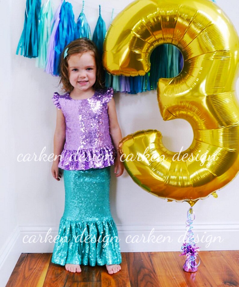 Puede incluir: Una ni&ntilde;a peque&ntilde;a con un disfraz de sirena de lentejuelas moradas y azul verdoso se encuentra frente a un gran globo de oro con el n&uacute;mero tres. El globo tiene una serpentina atada. La ni&ntilde;a sonr&iacute;e y mira a la c&aacute;mara. El fondo es una pared blanca con una guirnalda de borlas azules y moradas.