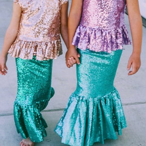 Op de afbeelding: Twee meisjes dragen bijpassende turquoise zeemeerminstaarten met paillettenrokken en -tops. E&eacute;n meisje draagt een roze paillettentop en de andere draagt een gouden paillettentop.