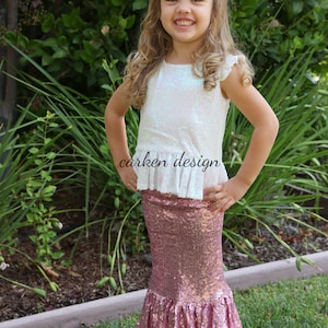 Puede incluir: Una ni&ntilde;a peque&ntilde;a lleva una blusa blanca con lentejuelas iridiscentes y un dobladillo con volantes y una falda de sirena de lentejuelas rosas. La falda tiene un dise&ntilde;o de cola de pez y est&aacute; hecha de tela brillante. La ni&ntilde;a sonr&iacute;e y est&aacute; de pie en un &aacute;rea cubierta de hierba.