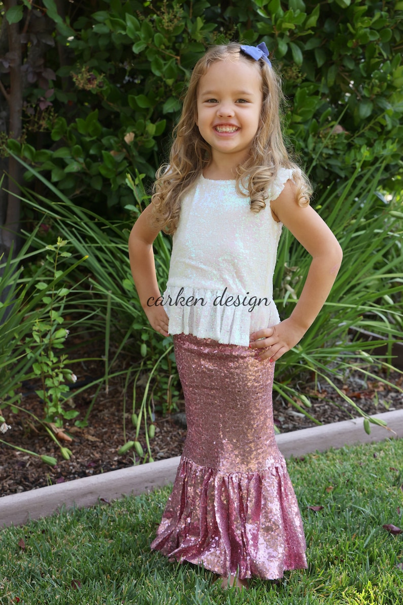 Puede incluir: Una ni&ntilde;a peque&ntilde;a lleva una blusa blanca con lentejuelas iridiscentes y un dobladillo con volantes y una falda de sirena de lentejuelas rosas. La falda tiene un dise&ntilde;o de cola de pez y est&aacute; hecha de tela brillante. La ni&ntilde;a sonr&iacute;e y est&aacute; de pie en un &aacute;rea cubierta de hierba.