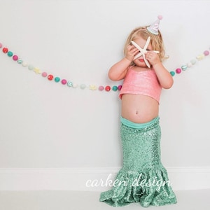 Op de afbeelding: Een jong kind draagt een roze crop top en een groene, met pailletten bezette zeemeerminnenstaartrok. Het kind houdt een witte zeester voor zijn gezicht en draagt een witte en zilveren feestmuts. Een kleurrijke kralenkrans hangt op de achtergrond.