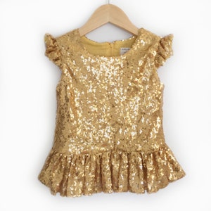 Puede incluir: Una blusa de lentejuelas doradas para ni&ntilde;as con mangas cortas con volantes y un dobladillo con volantes.