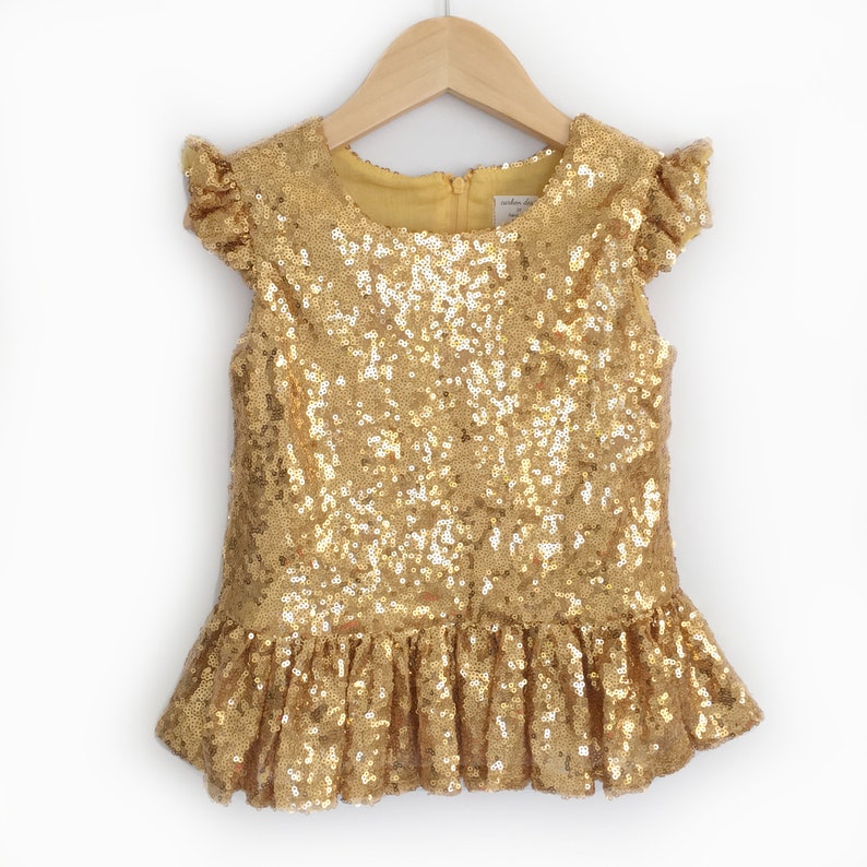 Puede incluir: Una blusa de lentejuelas doradas para ni&ntilde;as con mangas cortas con volantes y un dobladillo con volantes.