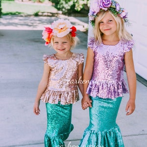 Puede incluir: Dos ni&ntilde;as con trajes de sirena con tops de lentejuelas y colas de sirena turquesa. Las ni&ntilde;as se toman de la mano y sonr&iacute;en. Una ni&ntilde;a lleva una corona de flores rosa y la otra lleva una corona de flores rojas y blancas.