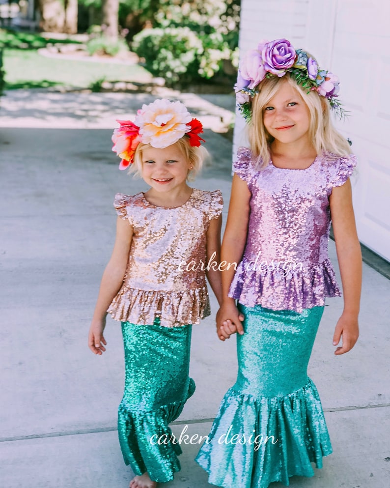 Puede incluir: Dos ni&ntilde;as con trajes de sirena con tops de lentejuelas y colas de sirena turquesa. Las ni&ntilde;as se toman de la mano y sonr&iacute;en. Una ni&ntilde;a lleva una corona de flores rosa y la otra lleva una corona de flores rojas y blancas.