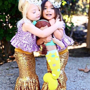 Op de afbeelding: Twee jonge meisjes dragen gouden glitter-zeemeerminstaarten en paarse tops met gouden kronen op hun hoofd. Ze houden een pop vast en knuffelen elkaar.