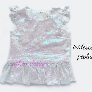 Puede incluir: Una blusa blanca de lentejuelas iridiscentes con un dobladillo de p&eacute;plum con volantes y mangas cortas. La blusa tiene un escote redondo y est&aacute; hecha de una tela suave y c&oacute;moda.