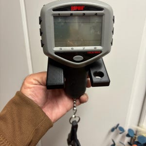 May include: A gray and black digital fish scale with the brand name "Rapala" in red. The scale has a digital display with numbers 1-8 and a power button. It is designed to measure up to 15 pounds or 6 kilograms.