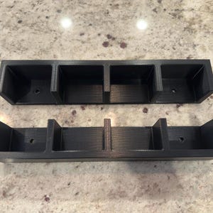 Può includere: Due organizzatori rettangolari neri con più scomparti. Ogni organizzatore ha una serie di aperture quadrate e un foro nella base di ogni scomparto. Gli organizzatori sono realizzati in un materiale nero opaco.