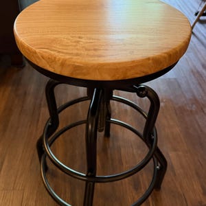 May include: A wooden adjustable bar stool with a round, light-colored seat and a black metal frame. The stool has a footrest and a mechanism for adjusting the height. The design is industrial and rustic, suitable for a kitchen or bar setting.