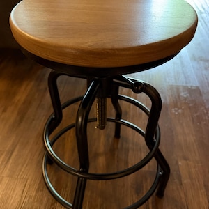 May include: A round wooden stool with a smooth, light brown finish on the seat. The base is constructed from black metal, featuring curved legs and a circular footrest. A central threaded metal mechanism allows for height adjustment of the seat.