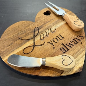 May include: A heart-shaped wooden cheese board with the words "Love you always" engraved on it. Includes a cheese fork and a cheese knife, both with wooden handles and heart-shaped cutouts. The board is made of light brown wood.