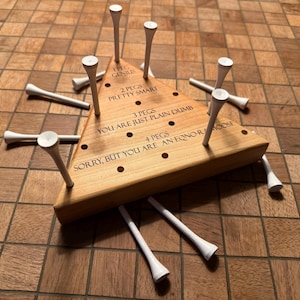 May include: A triangular wooden peg solitaire game board with holes and white golf tees. The board features engraved text: 1 PEG GENIUS, 2 PEGS PRETTY SMART, 3 PEGS YOU ARE JUST PLAIN DUMB, 4 PEGS SORRY, BUT YOU ARE AN EQNO-RAMOOSE. Several white tees are inserted.