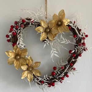 May include: A heart-shaped wreath made from brown twigs, white frosted branches, and red berries. The wreath is decorated with three gold glitter poinsettia flowers.