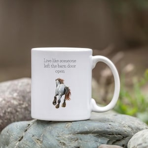 以下が含まれることがあります： 白いセラミックマグカップ。馬のイラストと「Live like someone left the barn door open」のテキスト入り。馬は灰色で、茶色のたてがみと尾があります。マグカップにはカーブしたハンドルが付いています。