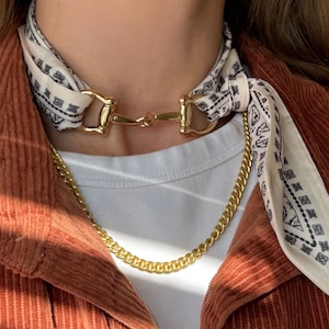 May include: A gold-colored necklace with a curb chain and a horse bit-style clasp. The necklace is paired with a cream-colored bandana with a black geometric pattern. The person is wearing a white shirt and a rust-colored corduroy jacket.