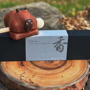 May include: A carved wooden capybara figurine with a light-colored wooden stick, resting on a long black rectangular box. The box features a gray label with the text Hainan Kyara Agarwood and a large Japanese character, plus a small capybara icon.