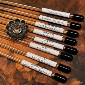May include: An assortment of incense sticks with white labels and black tips, featuring various scents like "Thai Agarwood" and "Red Cypress." A decorative bronze flower sits among the sticks. The background is a textured, brown surface.