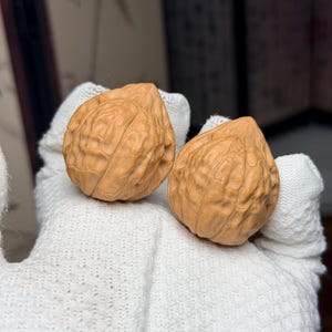Puede incluir: Dos cáscaras de nuez de color marrón claro con una parte superior puntiaguda y una superficie irregular profundamente surcada. Las cáscaras descansan sobre una superficie de tela blanca texturizada, mostrando un aspecto natural y leñoso.