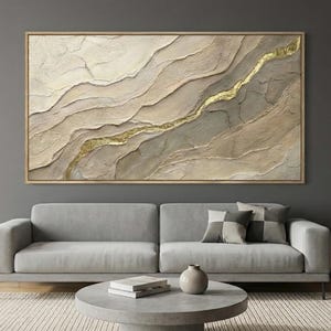 May include: Large abstract wall art with a textured surface in neutral tones of beige, grey, and gold. The artwork is framed in a light wood and hangs above a grey sofa. The room has a modern, minimalist design.
