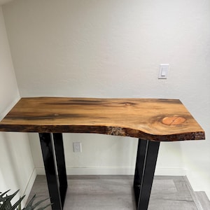 Puede incluir: Una consola de madera de borde vivo con un acabado de vetas naturales. La encimera está soportada por patas de metal negro. La mesa mide aproximadamente 122 cm de largo y 41 cm de ancho.