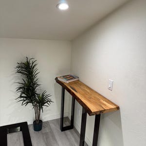 Puede incluir: Una consola de borde vivo con una tapa de madera natural y patas de metal negro. La mesa mide aproximadamente 152 cm de largo y tiene un libro encima. Una planta en maceta está a la izquierda.