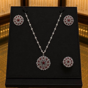 Peut inclure: Un ensemble de bijoux en argent présenté sur une surface noire. L'ensemble comprend un collier, des boucles d'oreilles et une bague, tous avec un motif circulaire orné de pierres rouges. Le collier a un pendentif avec un plus grand arrangement de pierres précieuses.
