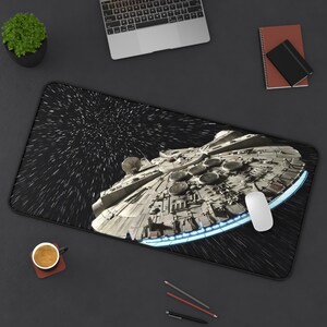 Może przedstawiać: Prostokątna podkładka na biurko z grafiką statku kosmicznego Millennium Falcon podróżującego przez przestrzeń kosmiczną na tle gwiazd. Podkładka ma czarną obwódkę i przedstawia statek w odcieniach szarości z niebieskim światłem silnika.