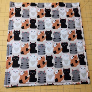 May include: A square, quilted fabric with a repeating pattern of cartoon cats. The cats are in various colors, including black, gray, orange, and white, with heart-shaped details. The background is a light beige.
