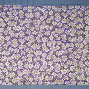 Può includere: Un tessuto rettangolare con un motivo ripetuto di margherite bianche con centri gialli su sfondo viola. Le margherite sono uniformemente distanziate, creando un fitto disegno floreale. Adatto per artigianato o decorazioni per la casa.