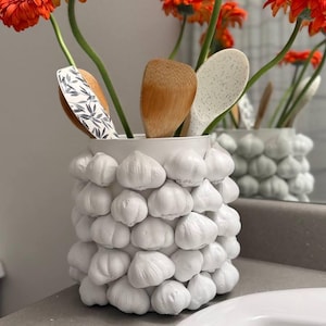 Könnte beinhalten: Ein weißer Keramik-Utensilienhalter, verziert mit Knoblauchzehen, mit Holzlöffeln und orangefarbenen Gerbera-Gänseblümchen. Der Halter steht auf einer grauen Arbeitsplatte in der Nähe eines weißen Waschbeckens.