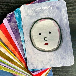 May include: A deck of colourful cards with rounded corners, fanned out to display the top card. The top card has a watercolour illustration of a face and the text "I feel empty."