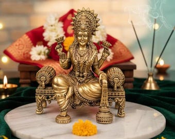 May include: A gold-coloured statue of a seated deity, with detailed jewellery and a halo, holding flowers. The statue is on a decorative seat, placed on a white marble surface. Incense sticks and flowers are in the background.