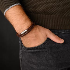 May include: A brown leather bracelet with a braided design. The bracelet wraps around the wrist twice and features a silver clasp. The image shows a close-up of the bracelet on a person's wrist.