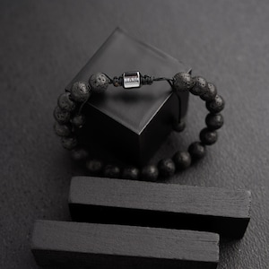 Peut inclure: Bracelet de perles en pierre de lave noire avec un accent en métal argenté. Le bracelet est monté sur un cordon noir et est présenté sur un bloc géométrique noir et deux blocs rectangulaires noirs.