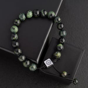 May include: A beaded bracelet featuring dark green and black speckled beads. The bracelet is strung on a black cord and has a silver-colored square accent piece with a design. The bracelet is displayed on a black surface.