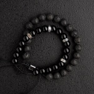 May include: Two beaded bracelets. One bracelet features black onyx beads and silver accents. The other bracelet has dark gray lava stone beads. Both bracelets are on a black surface.