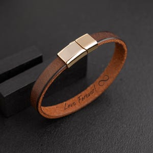May include: Brown leather bracelet with a gold-colored clasp. The bracelet has the words "Love Forever!" and an infinity symbol engraved on it. The bracelet is a simple, elegant design.