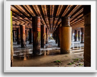 May include: A framed photograph captures the underside of a pier, showcasing weathered wooden pilings and beams. The perspective draws the eye down a corridor of columns, with sunlight filtering through, creating a contrast of light and shadow. The scene evokes a sense of age and the sea.