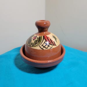 May include: A brown ceramic tagine with a lid. The lid features a floral pattern in red, green, and black on a cream base. The tagine rests on a matching base, creating a cohesive design.