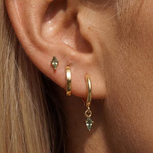 Peut inclure: Gros plan sur une oreille ornée de trois boucles d'oreilles dorées. Une petite puce avec une pierre verte, un simple anneau doré et un anneau doré avec une pierre verte pendante. Les bijoux sont mis en valeur sur l'oreille et les cheveux blonds.