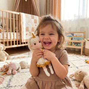 May include: A smiling child holds a handmade crochet doll with a unicorn horn. The doll has a white dress with yellow trim and white boots. The background includes a crib, toys, and a patterned rug.